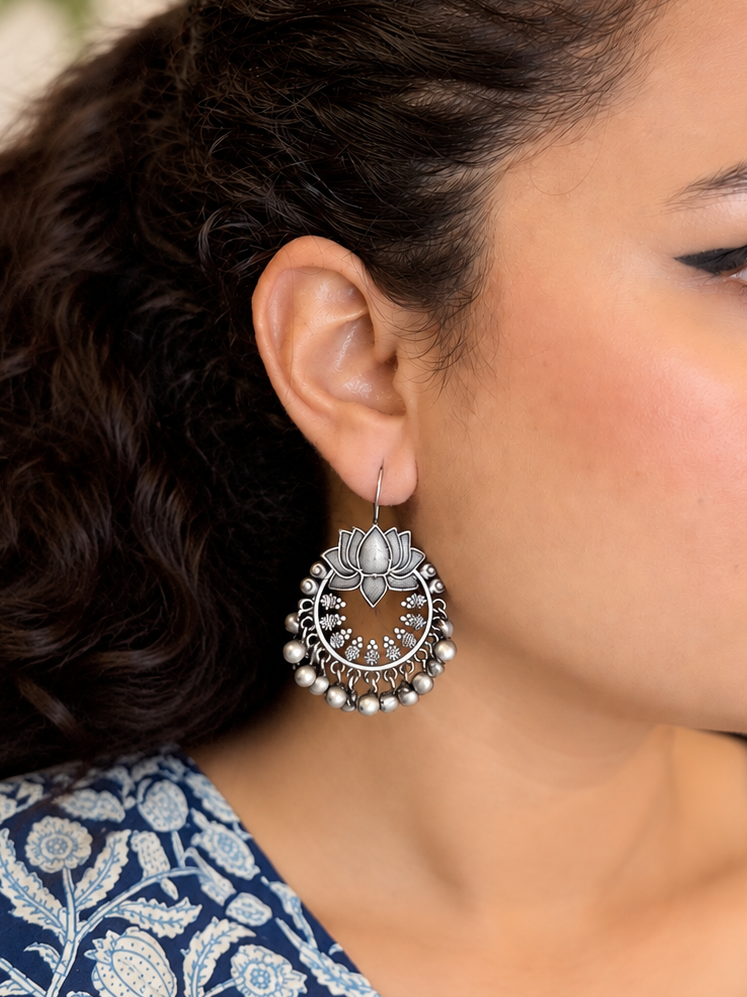 A close-up of a woman's ear wearing a detailed silver-toned huggie hoop earring with a lotus flower motif and dangling pearl-like beads.